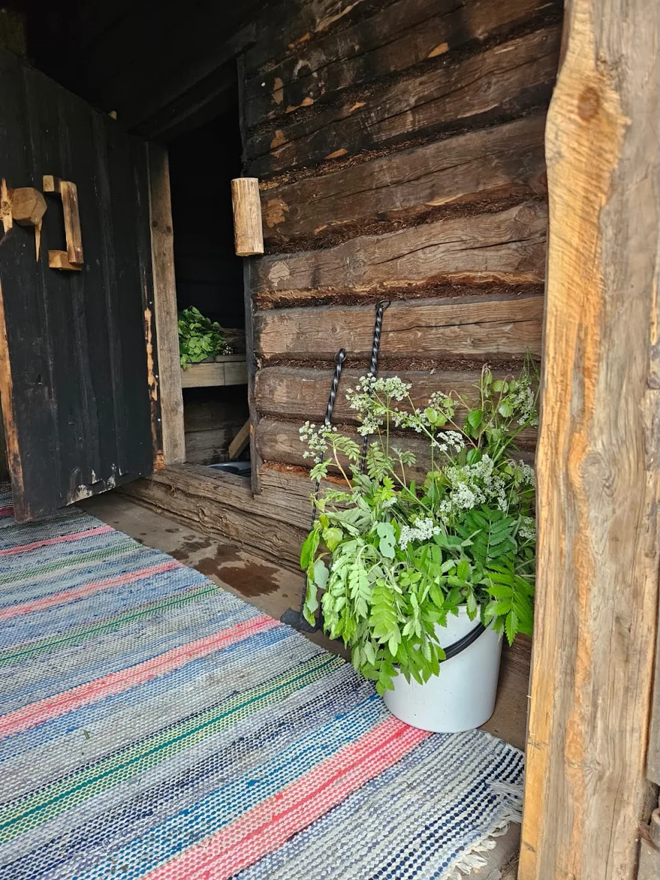 Perinnesaunan tumma hirsiseinä, avoin ovi ja räsymatto lattialla.